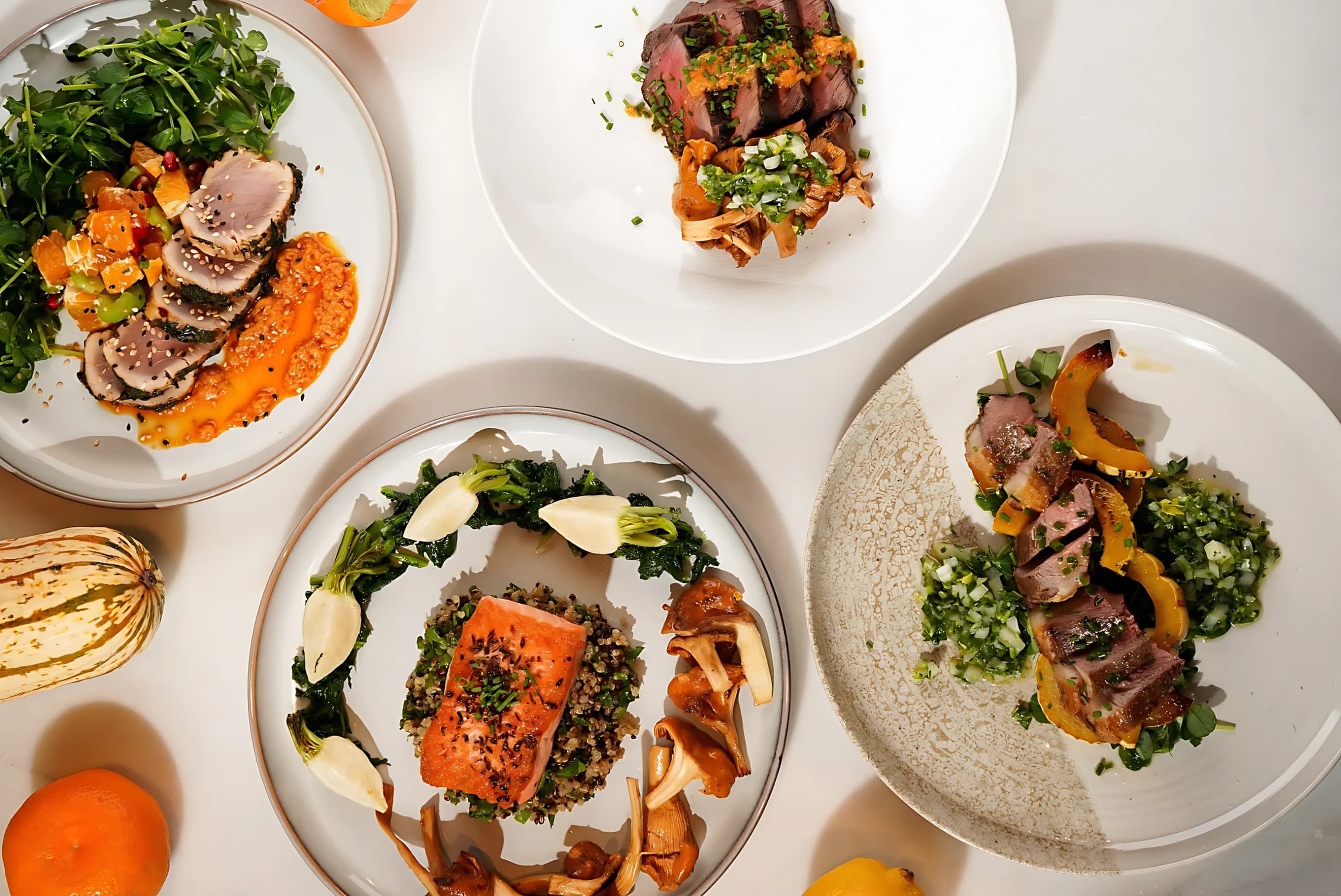 Plates of salmon, duck and greens laid out on a table with citrus and squash